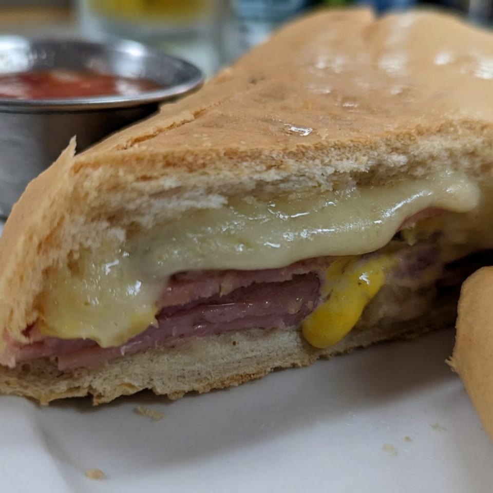 Close-up of a toasted steak sandwich with melted cheese and mustard, served with tomato dipping sauce.