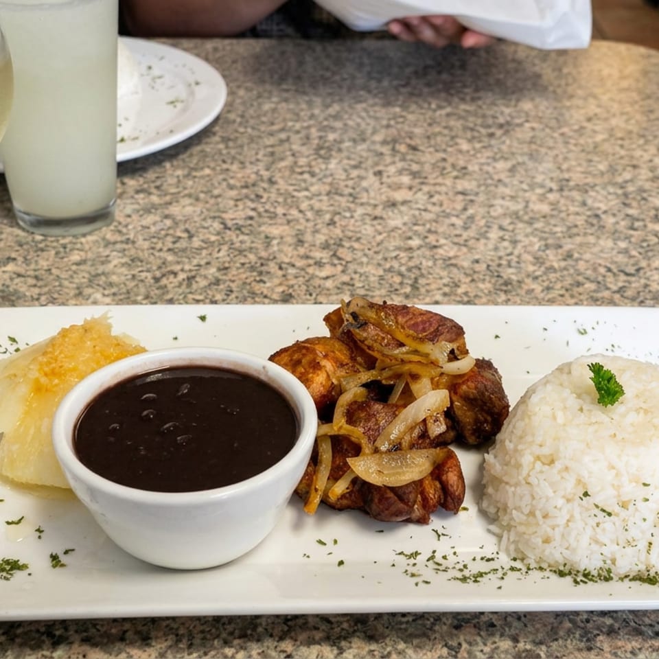 Plate of grilled meat with sautéed onions, white rice, mashed plantains, and black beans on a restaurant table.
